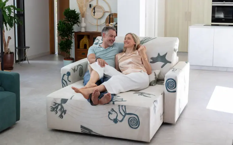 Pareja sentada en un sofá chaise longue tapizado en tela Quetzal, un tejido claro con motivos decorativos en tonos azules y grises, en un salón moderno y luminoso.
