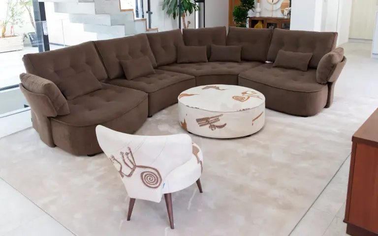 Silla y puff tapizados en tela Quetzal, un tejido claro con motivos decorativos en tonos tierra, combinados con un sofá modular marrón en un salón moderno y luminoso.