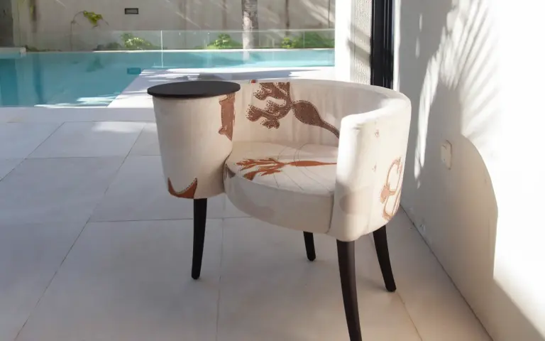 Sillón La Caracola tapizado en tela Quetzal, un tejido claro con motivos decorativos en tonos tierra, junto a una piscina en un ambiente luminoso y contemporáneo.
