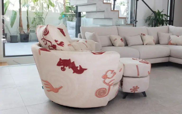 Sillón y puff tapizados en tela Quetzal, un tejido claro con motivos decorativos en tonos rojizos y rosados, combinados con un sofá modular claro en un salón moderno y luminoso.